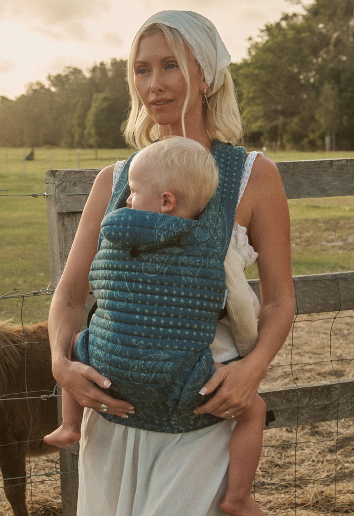 Front-facing view of Armadillo Baby Carrier in Vintage Blue worn with sleeping baby at golden hour farm setting